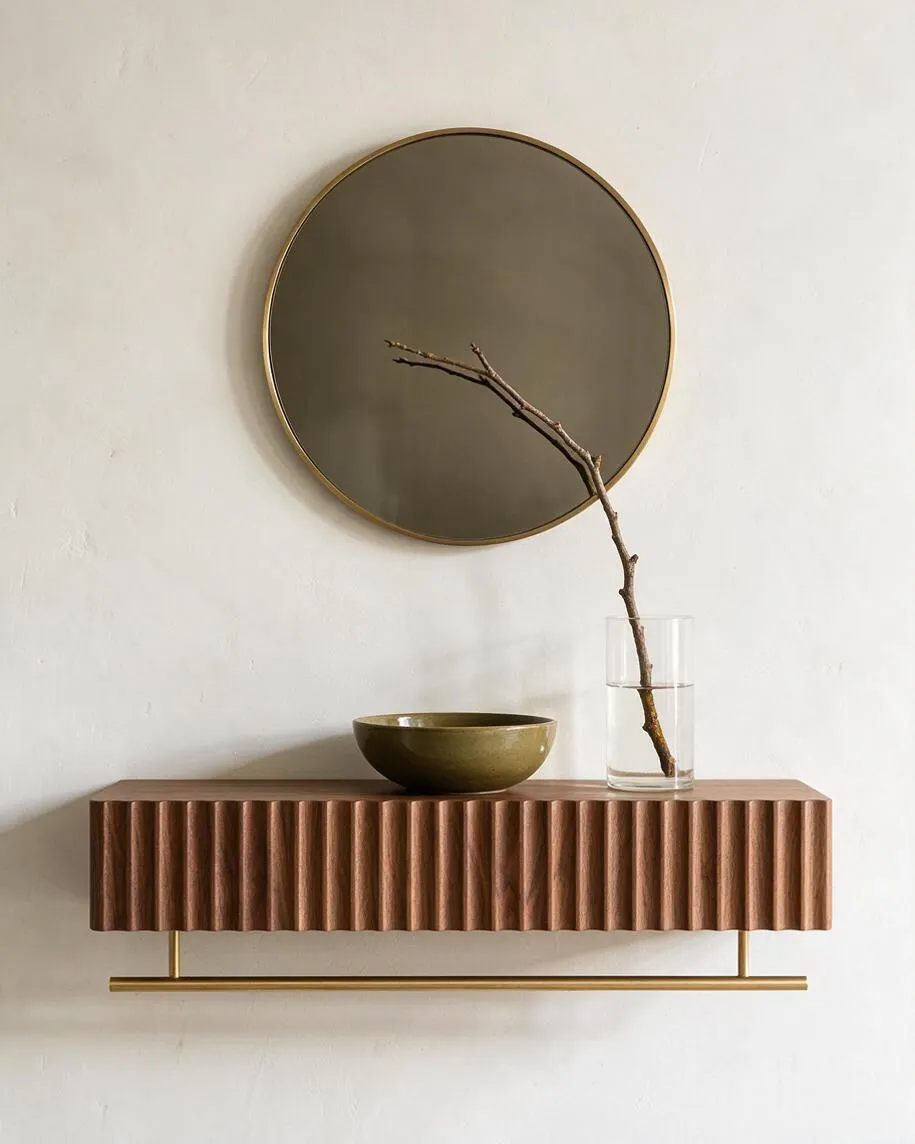 A modern wall vignette against a white plaster wall features a round dark mirror with a slim brass frame above a floating fluted wood shelf. On the shelf sit an olive-green bowl and a clear glass vase holding a single bare branch, with a brass support bar below.