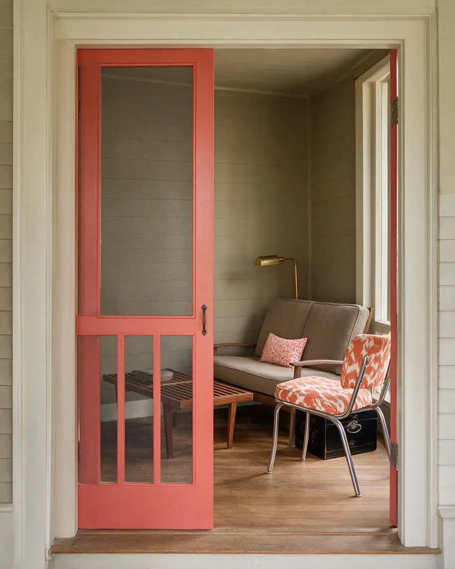 A front porch seating nook viewed through a coral-painted screen door, featuring a taupe loveseat, a patterned orange-and-white chair, a slatted wood coffee table, and a brass reading lamp. The porch has beige walls, a wood floor, and layered vintage-inspired accents.