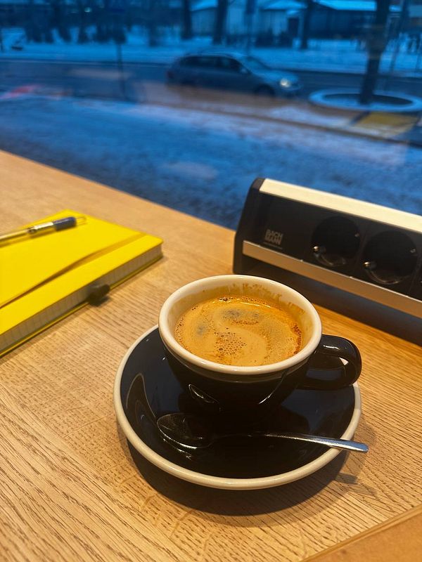 A cup of coffee sits on a wooden table next to a yellow notebook and a pen, with a snowy street visible through the window.