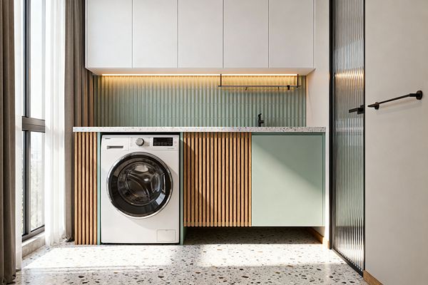 A compact contemporary laundry features a front-loading washer beneath a speckled terrazzo countertop, with white flat-panel wall cabinets above. The backsplash is sage green vertical fluted tile lit by a warm LED strip; base cabinets combine a mint-green slab door and a slatted oak panel, with a matte-black faucet and hanging rail, terrazzo flooring, a tall window with layered curtains to the left, and a reeded-glass door with black frame to the right.