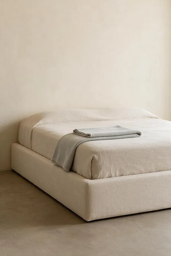 A minimalist bedroom features a low platform bed upholstered in soft beige fabric against a plain warm wall. The bed is dressed in neutral bedding with a folded light gray throw placed neatly near the foot, creating a calm, understated look.