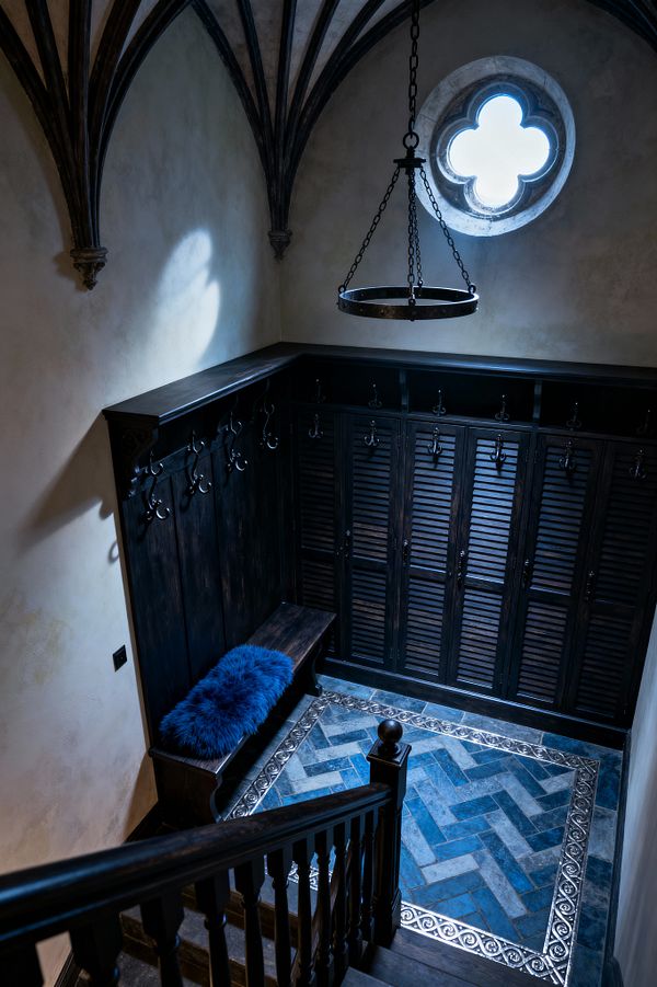 A Gothic-inspired mudroom at the base of a staircase features a ribbed vaulted ceiling, a round quatrefoil window, and a black metal ring chandelier. Dark-stained wood lockers with louvered doors and vertical paneling line two walls with scrolled wrought-iron hooks and a bench; the floor is blue and gray herringbone tile framed by a pewter scroll border, and a cobalt faux-fur cushion adds a soft accent.