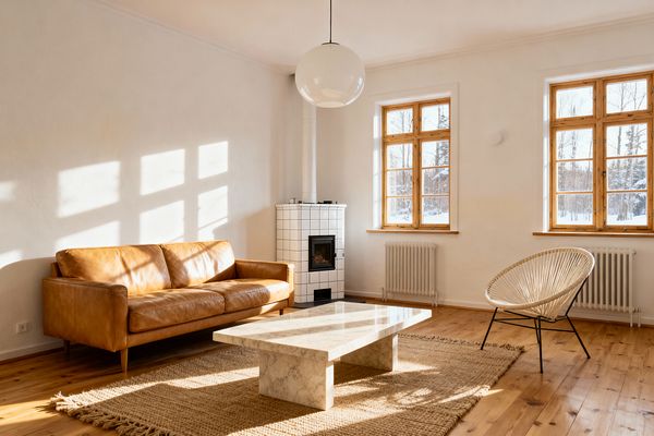 A bright Scandinavian living room with white plaster walls and natural wood floors. On the left, a cognac leather sofa faces a low rectangular white marble coffee table on a chunky jute rug; to the right, a woven cord lounge chair with black metal legs sits near two wood-framed windows, while a white square-tiled stove with a small black firebox stands in the corner beneath a round white globe pendant.