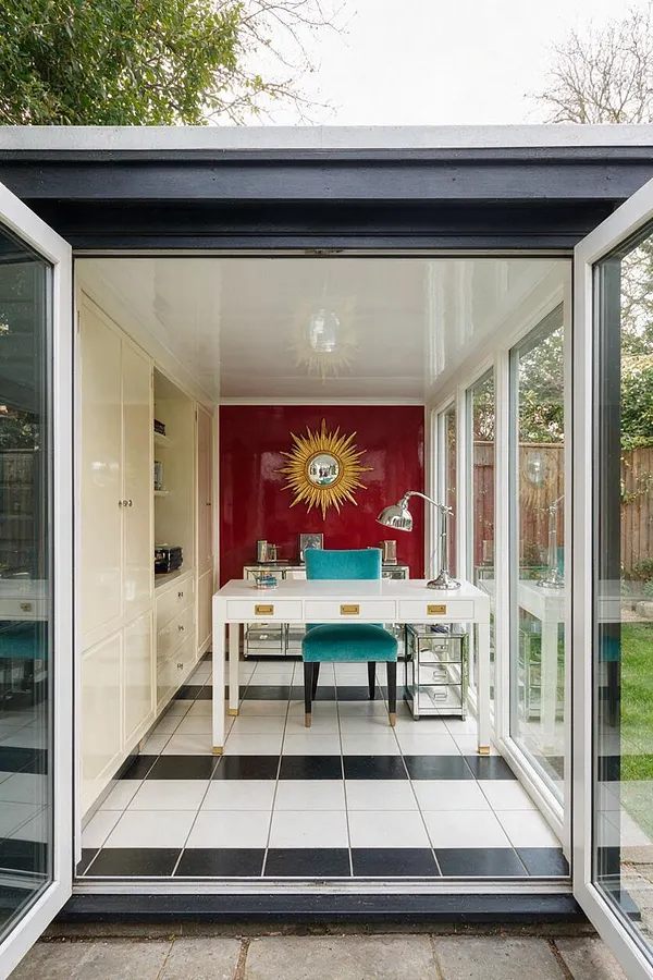 A compact backyard studio with wide open glass doors, a glossy white ceiling, and a black-and-white tiled floor. Inside, a white desk, turquoise chair, mirrored accents, and a red feature wall with a gold sunburst mirror create a bold Hollywood Regency look.