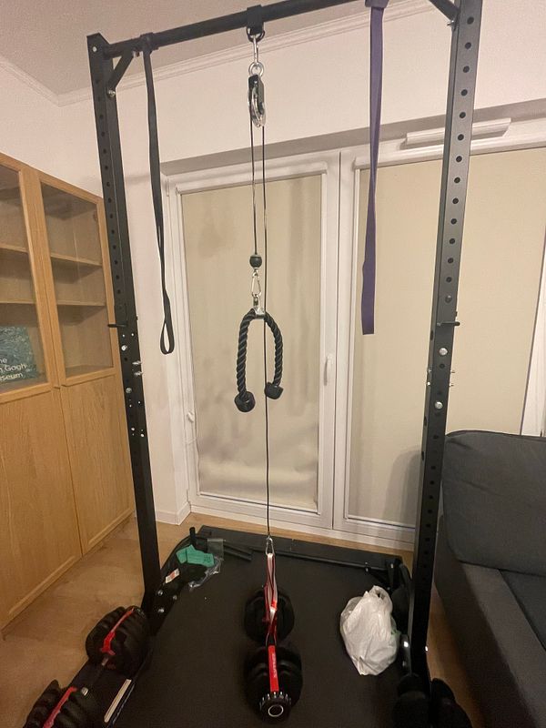 Home gym setup with a black power rack outfitted with a cable high pulley system and various accessories.