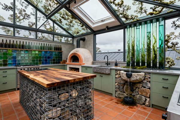 A greenhouse-style kitchen with a steel-framed glass roof and skylight features sage green base cabinets, concrete countertops, and a terracotta tile floor. A dome wood-fired oven, a gabion island topped with a thick live-edge wood slab, a blue–green recycled glass tile backsplash with a shelf of bottles, a standard stone sink plus a cascading stone basin fountain, and a row of tall green liquid-filled tubes with plantlike growth create a rugged, self-reliant aesthetic.