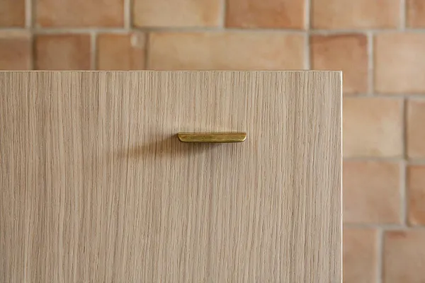 A close-up view of a light oak cabinet front with subtle vertical grain and a small brushed brass pull centered near the top. Behind it, a softly blurred terracotta tile wall adds warm earthy texture and color.