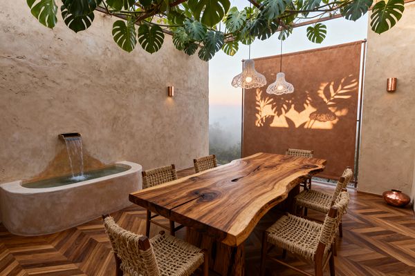 A biophilic dining room with warm plaster walls features a live-edge hardwood table and six wooden chairs with jute-woven seats on herringbone wood flooring. Overhead, a trellis supports large monstera leaves and three white woven pendant lights. A plaster trough fountain with a metal spout flows along one wall, while a tan canvas screen at the opening captures leafy shadows with a misty landscape visible beyond.