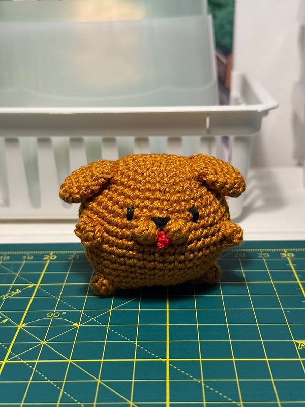 A crocheted critter resembling a small, round dog sits on a cutting mat.