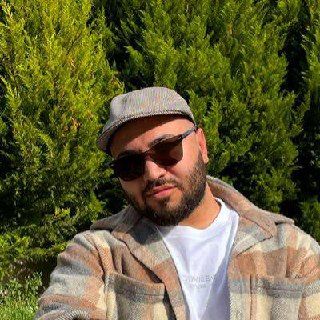A man poses outdoors in front of lush green foliage.