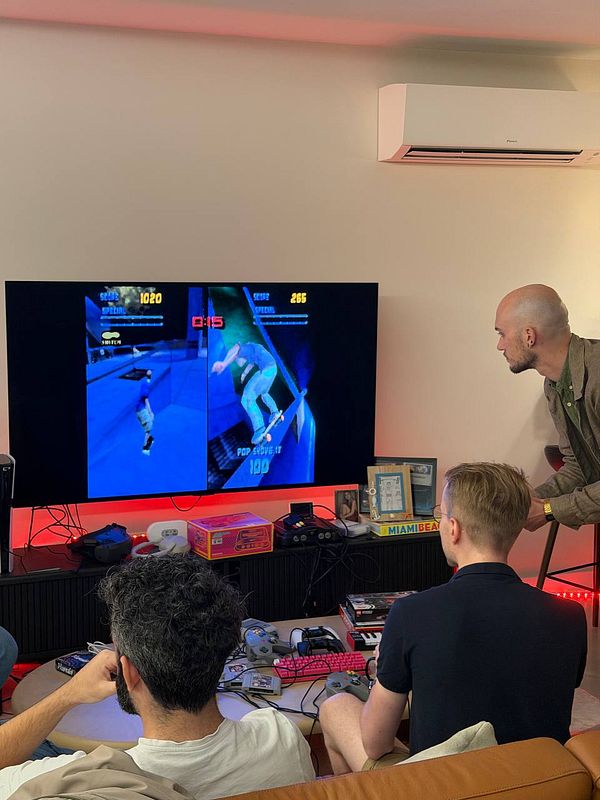 Three men playing a split-screen skateboarding video game in a cozy indoor setting with retro gaming consoles.