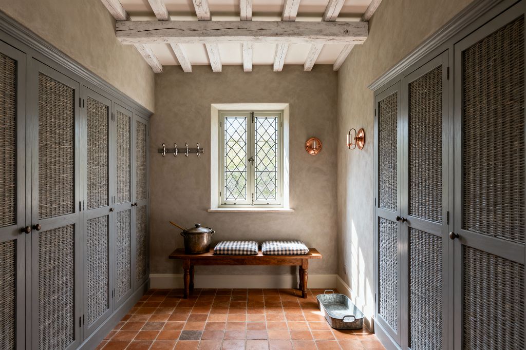 A narrow French country mudroom features symmetrical gray built-ins with woven rattan doors on both sides. Centered under a small leaded-glass casement window is a wood bench topped with two gingham cushions; rustic terracotta tiles, limewashed plaster walls, whitewashed ceiling beams, copper wall sconces, a row of metal hooks, and a galvanized boot tray complete the warm, utilitarian space.