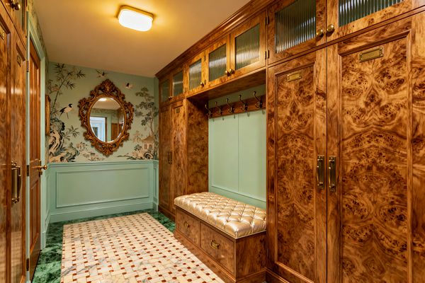 A vintage-glam mudroom with symmetrical built-in lockers clad in honey-toned burl wood and ribbed glass uppers, finished with polished brass pulls, label plates, and coat hooks. A mint-painted alcove holds a tufted champagne leather bench with drawers below, while mint wainscoting meets chinoiserie wallpaper with birds and an ornate gilded oval mirror; the floor is a marble mosaic with terracotta dots framed by a deep green marble border beneath a warm flush-mount light.