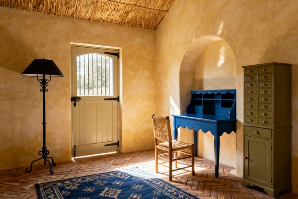 A sunlit French Country home office with limewashed ochre walls and a straw-thatch ceiling features a cream Dutch door with black iron hardware opening onto herringbone terracotta brick floors. A cobalt blue scalloped writing desk tucked into an arched niche pairs with a woven rush-seat chair, while an olive apothecary cabinet, blue patterned rug, and wrought-iron floor lamp create a warm, rustic workspace.