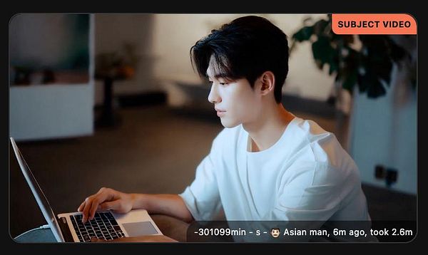 A young Asian man is focused on his laptop in a modern indoor setting.