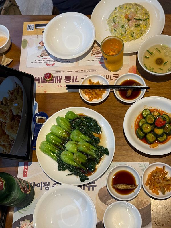A table set with various plates of Asian dishes including bok choy with sauce, shrimp fried rice, pickled cucumber, soup, dipping sauces, and a glass of tea accompanied by chopsticks and a tablet displaying food.
