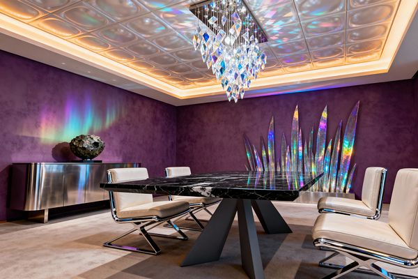 A dramatic dining room with deep purple textured walls features a black marble dining table on angular gray pedestals, surrounded by ivory leather chairs with chrome bases. A faceted crystal chandelier and an iridescent blade-like wall sculpture project rainbow reflections onto a backlit coffered ceiling, while a stainless steel credenza topped with a large geode and a soft taupe carpet complete the polished, futuristic look.