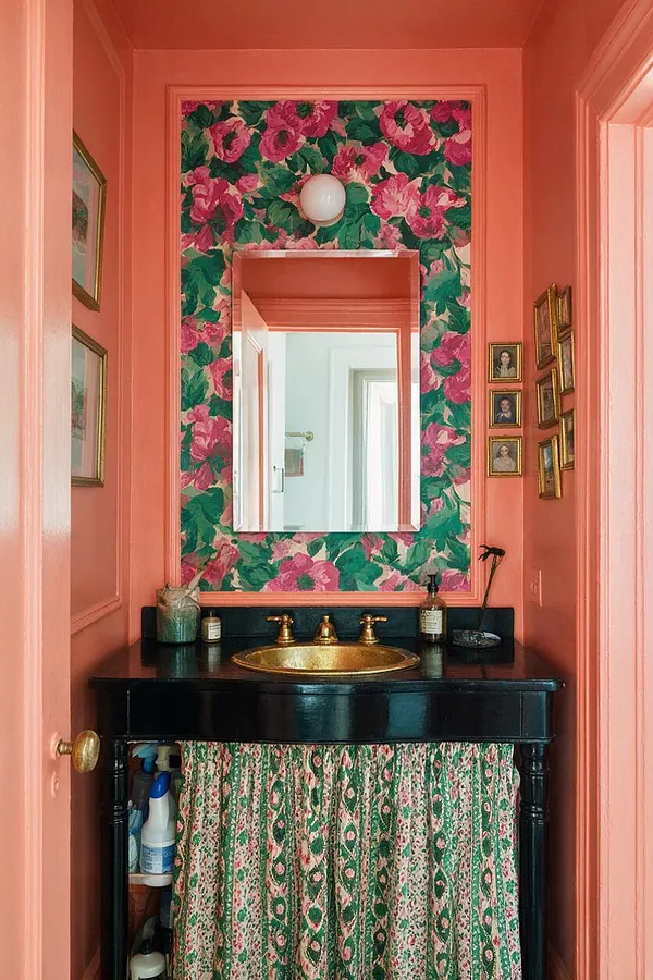 A narrow powder room painted in coral pink with a large floral wallpaper panel framing a rectangular mirror above a black vanity. The vanity has a gold faucet and sink, patterned fabric curtain below, and small framed portraits and decor clustered on the side walls.