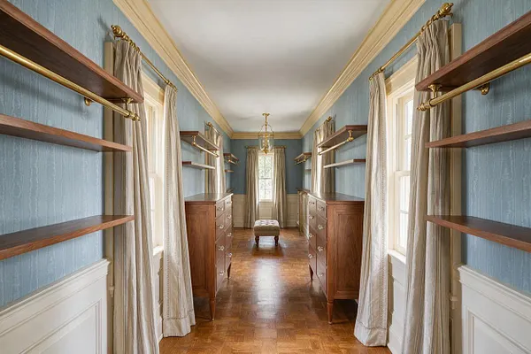 A long traditional walk-in closet with blue textured walls, white crown molding, parquet wood flooring, and symmetrical walnut shelving along both sides. Cream curtains frame the windows, while matching wood dressers with brass hardware and a small upholstered ottoman sit along the center axis.