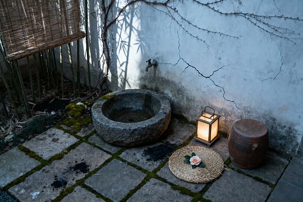 A small Wabi-Sabi patio corner features a rough stone water basin beneath a simple wall faucet, set on large stone pavers threaded with moss. A cracked plaster wall with bare climbing vines forms the backdrop, while bamboo canes and a woven reed screen cast soft shadows. A glowing wood-framed rice paper lantern sits beside a weathered terracotta jar and a round straw mat topped with a single pink blossom, creating a serene, timeworn scene.