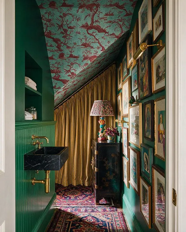 A narrow powder room with emerald green walls, a black marble wall-mounted sink, and brass plumbing and sconce. The ceiling is covered in turquoise and burgundy botanical wallpaper, one wall is filled with framed art, and gold drapery and patterned rugs add a richly layered maximalist look.
