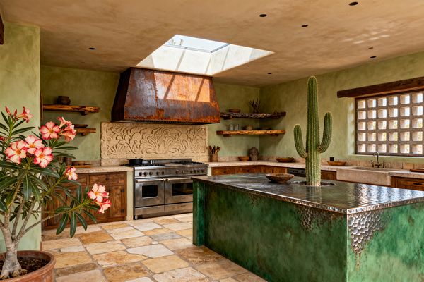 Southwestern kitchen with sage plaster walls, a large skylight, and a verdigris hammered-copper island featuring a tall cactus planter. A rusted metal range hood sits over a stainless steel range, backed by a carved limestone backsplash. Rough-hewn wood shelves and knotty wood base cabinets frame stone countertops. Tumbled travertine floor tiles, a thick stone farmhouse sink, and a breezeblock window complete the earthy, sunlit space.