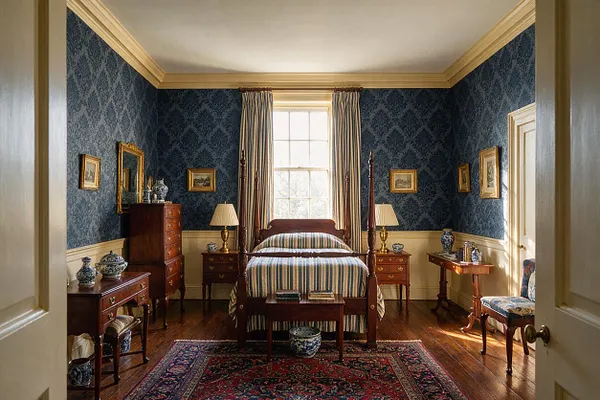 A traditional master bedroom with deep blue damask wallpaper, cream trim, and a central four-poster bed dressed in striped bedding. The room includes matching wooden nightstands, antique-style chests, framed artwork, a patterned red rug, and daylight coming through a tall window with draped curtains.