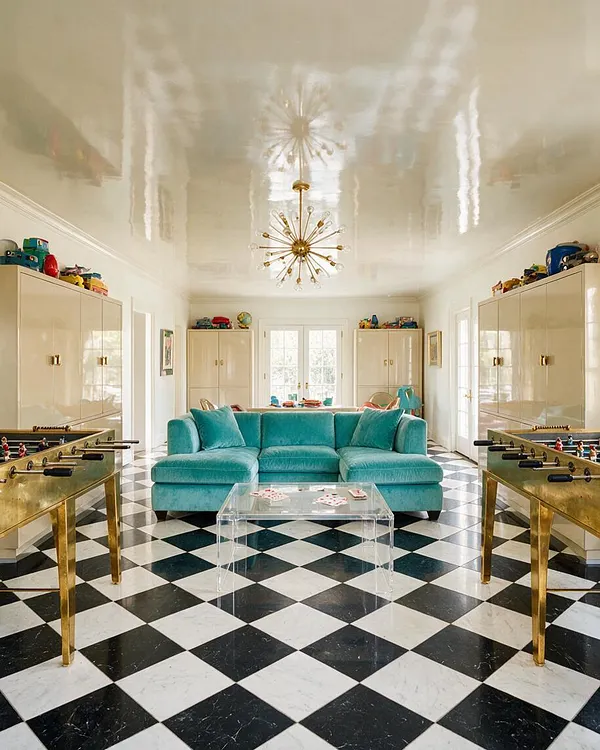 A bright game room in Hollywood Regency style features a black-and-white diamond marble floor, a glossy cream ceiling, and a teal velvet sectional centered in the room. Brass foosball tables, mirrored cabinetry, and a gold starburst chandelier add glamorous, playful contrast.