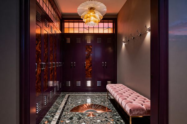 An Art Deco mudroom with deep plum locker-style cabinets featuring burled-wood panels, louvered vents, and polished chrome pulls. A green-black terrazzo floor with a copper fan inlay sits beneath a tiered scalloped glass pendant; an amber-and-rose stained-glass transom glows above, while a long blush velvet tufted bench on brass legs lines the right wall beneath a row of chrome coat hooks and warm gray walls.
