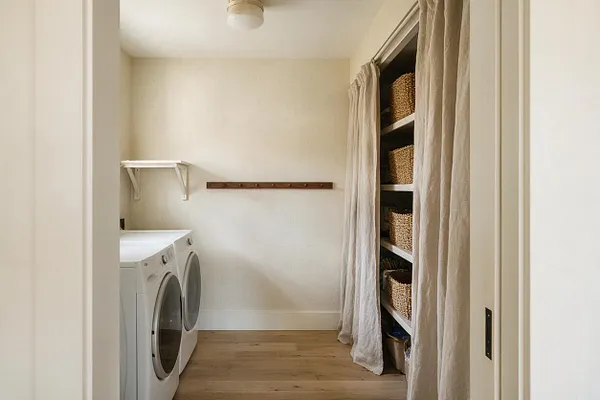 A narrow laundry room with a white front-loading washer and dryer on the left, open shelving with woven baskets on the right, and a simple wooden wall-mounted drying rack centered on the back wall. Soft beige curtains partially conceal the shelving, while warm wood flooring and cream walls create a calm, organic modern look.