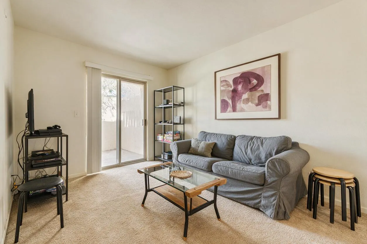 A simple modern game room with beige carpet, off-white walls, and a gray upholstered sofa beneath abstract wall art. The room includes a glass-top coffee table, a small TV on a black metal stand, open shelving, and stacked stools near the corner, with sliding glass doors bringing in daylight.