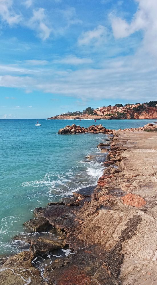 A sandy beach with gentle waves and partly cloudy skies, viewed from a rocky edge with some plants.