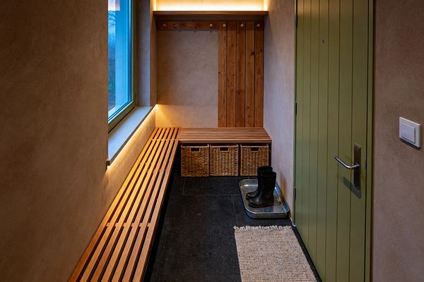 Compact mudroom with warm beige plaster walls and a sage green, vertical‑plank door. A long slatted wood bench runs beneath a window and turns into a corner seat above three woven baskets. Vertical timber boards with simple pegs form a hanging area. Concealed LED strips glow under the windowsill and shelf, lighting dark slate flooring. A jute runner and a galvanized tray with black boots underscore functional Scandinavian simplicity.