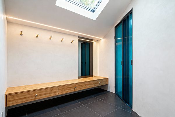 A modern mudroom with smooth white walls, a floating light-wood bench with three drawers, and a full-height mirror at the end. A recessed ceiling LED and a square skylight illuminate matte charcoal tiles; five brass hooks line the wall above the bench, and black-framed sliding doors with ribbed teal glass complete the bright, uncluttered space.