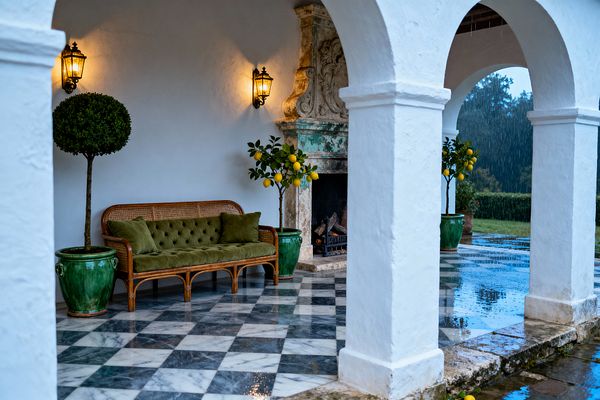 A covered patio with white stucco arches frames a checkerboard marble floor, a carved stone fireplace with green patina, and a rattan settee with tufted olive velvet cushions. Warm wrought-iron wall lanterns glow above potted topiary and lemon trees in emerald ceramic jars, while rain glistens on the floor and a garden landscape stretches beyond.