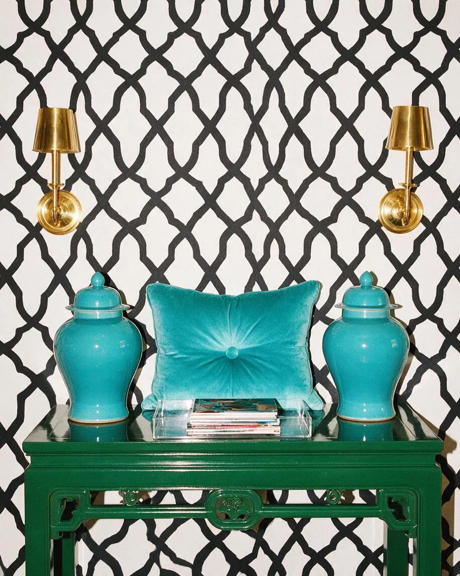 A Hollywood Regency-style vignette with a glossy emerald green console table against a white wall covered in bold black lattice-pattern wallpaper. Two brass wall sconces flank a turquoise velvet pillow, with matching turquoise ceramic ginger jars and a clear acrylic tray of magazines arranged on top.