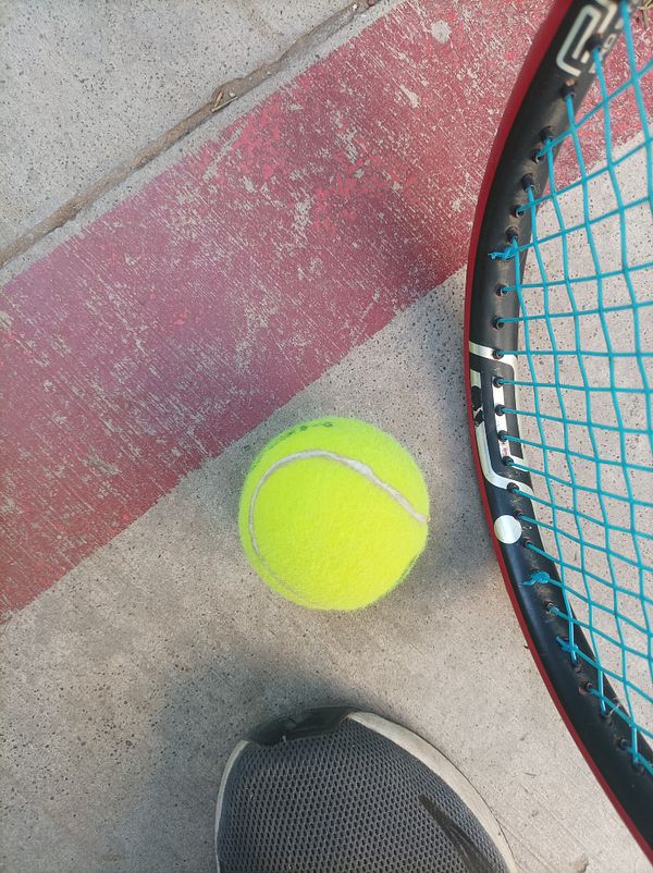 A tennis ball is positioned on the ground next to a tennis racket and a sneaker.