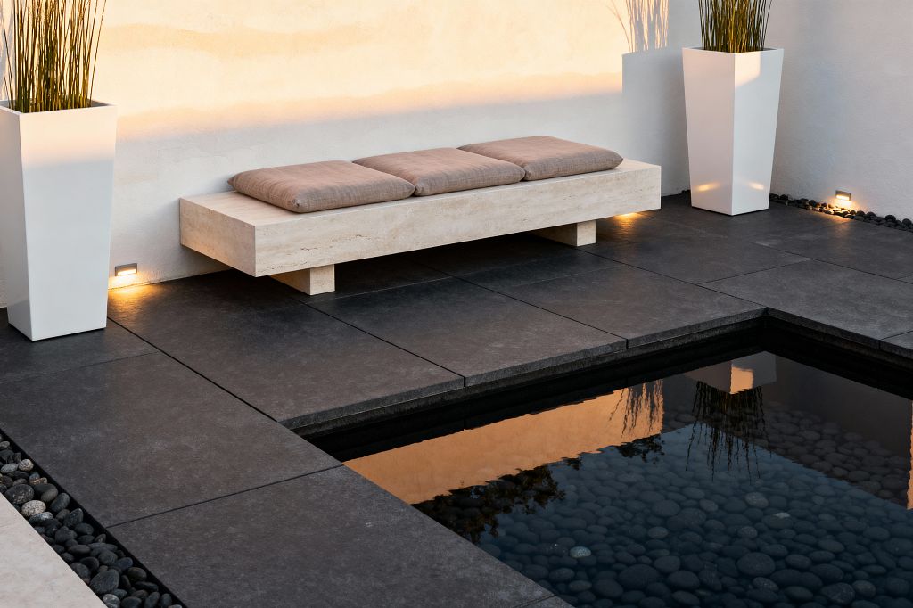 A minimalist patio features a light travertine bench with three taupe cushions set against a warm white stucco wall. Charcoal large-format pavers frame a shallow reflecting pool filled with smooth dark river pebbles, while two tall matte white planters with slender grasses flank the scene. Discreet recessed LED step lights wash the wall and floor with a soft golden glow, highlighting the clean lines and serene, uncluttered composition.