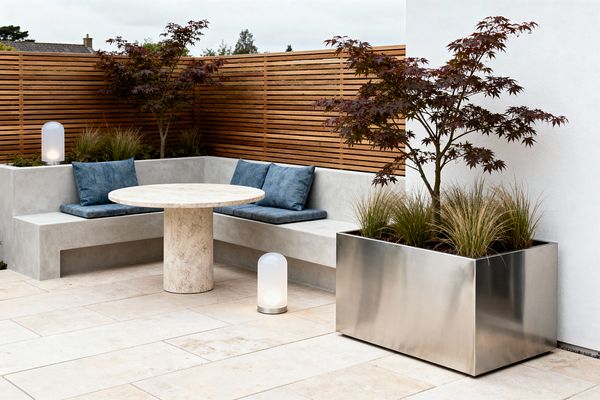 Contemporary outdoor seating area with an L-shaped light gray concrete bench and blue seat cushions surrounding a round travertine pedestal table on large cream limestone pavers, enclosed by a warm cedar slatted privacy fence. A brushed stainless-steel planter holds a burgundy-leaved Japanese maple and ornamental grasses, echoed by planting behind the bench, while two portable frosted-glass lanterns provide soft, low lighting.