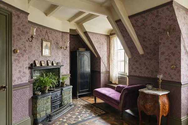 A Victorian-style studio apartment with floral lavender wallpaper, exposed cream ceiling beams, and a patterned tile floor. The room includes an ornate green-tiled fireplace, a deep purple chaise lounge, a black armoire, and a carved wooden side table near a sunlit window with lace curtains.