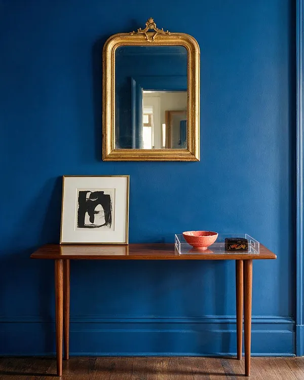 A vivid cobalt-blue wall anchors a narrow console vignette with an ornate gold-framed mirror above a slim wooden table. On the table sit a framed black-and-white abstract print, a coral bowl in a clear acrylic tray, and a small decorative box, creating an eclectic mix of classic and modern pieces.