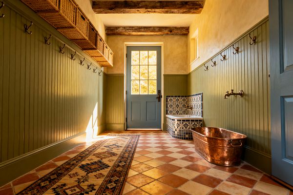 A farmhouse mudroom with sage-green beadboard walls lined with brass hooks, a row of woven baskets overhead, and a dusty blue door with divided lights at the end. Terracotta and cream checkerboard tiles span the floor with a patterned runner. A curved bench tiled in blue-and-white ceramic and a patinated copper tub sit to the right beneath exposed wood beams, all lit by warm natural light.