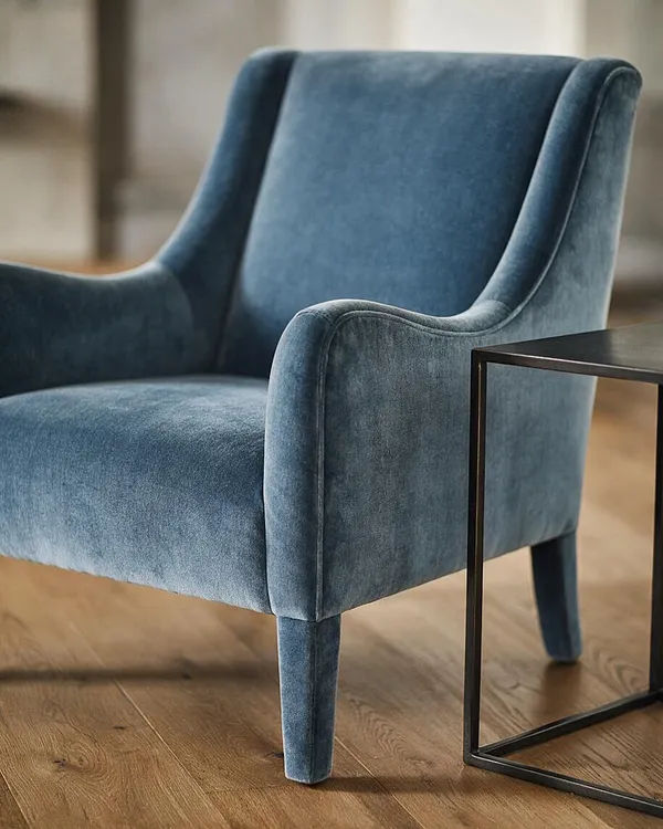 A transitional-style armchair in blue velvet with a gently curved back, padded arms, and tapered legs sits on a light wood floor. A slim black metal side table with a square frame is positioned beside it, creating a refined, modern-classic look.