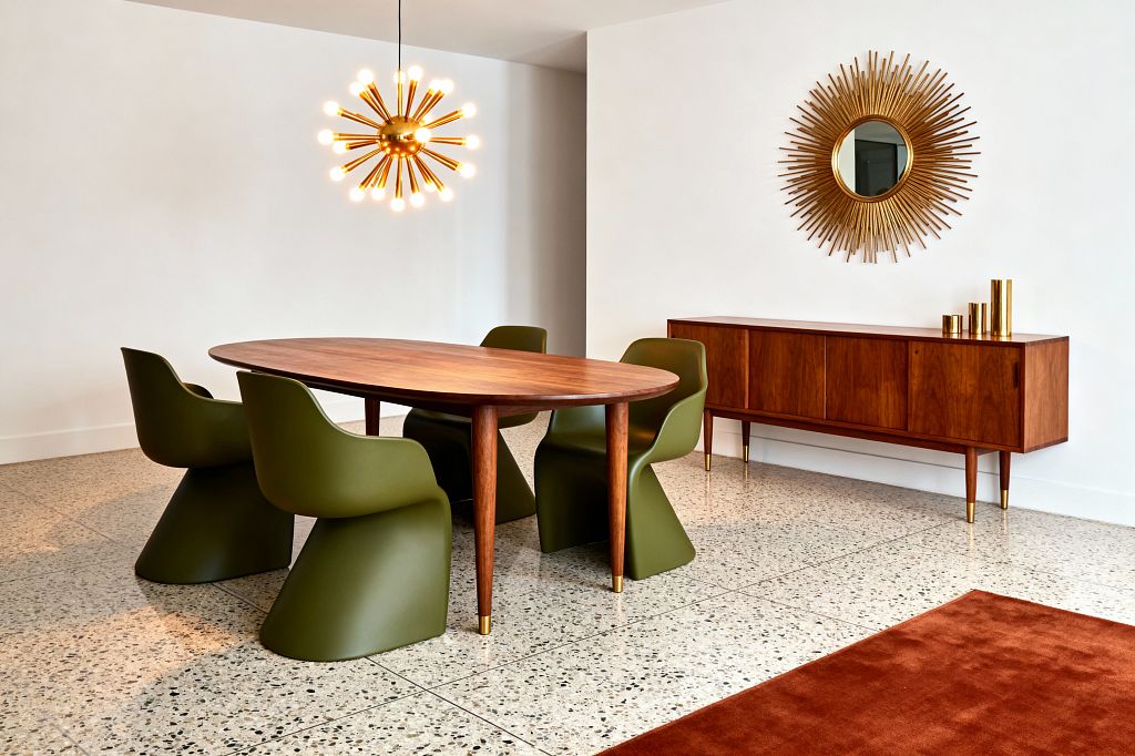A mid-century modern dining room features an oval walnut table with tapered legs and brass-capped feet, surrounded by five olive green molded chairs. A brass sputnik chandelier hangs above while a long walnut credenza with brass-tipped legs sits against white walls, topped by a gold sunburst mirror and brass cylinder vases, on speckled terrazzo flooring with a rust area rug in the foreground.