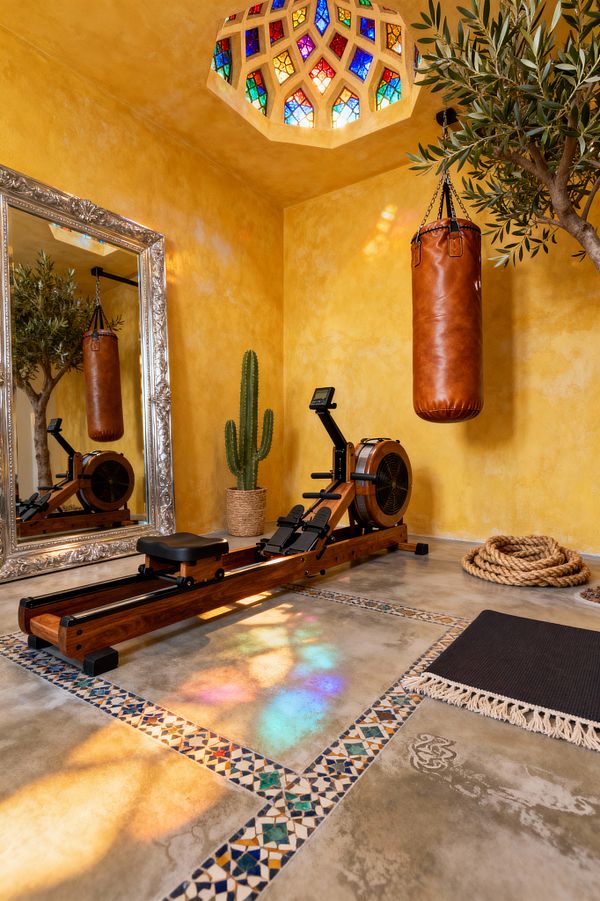 A Moroccan-style home gym with golden tadelakt plaster walls and an ornate silver-framed mirror. A solid-wood rowing machine sits on polished concrete bordered by blue-and-white zellige, facing a caramel leather hanging punching bag; coiled battle ropes, a black fringed mat, and a potted cactus rest nearby, while a stained-glass ceiling dome throws colorful light onto the floor beside an indoor olive tree.