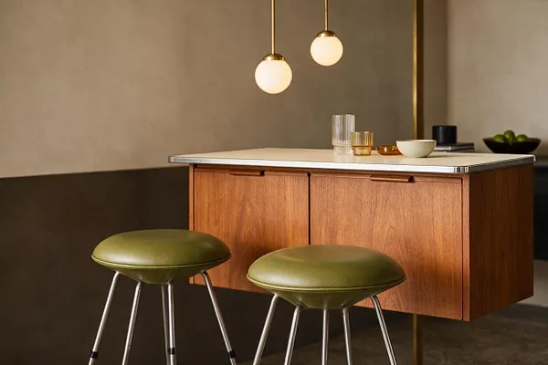 A Mid-Century Modern bar vignette with a floating walnut cabinet topped by a pale countertop, paired with two olive green cushioned stools on slender metal legs. Two globe pendant lights hang above, and small glassware and bowls sit on the counter against warm neutral walls.