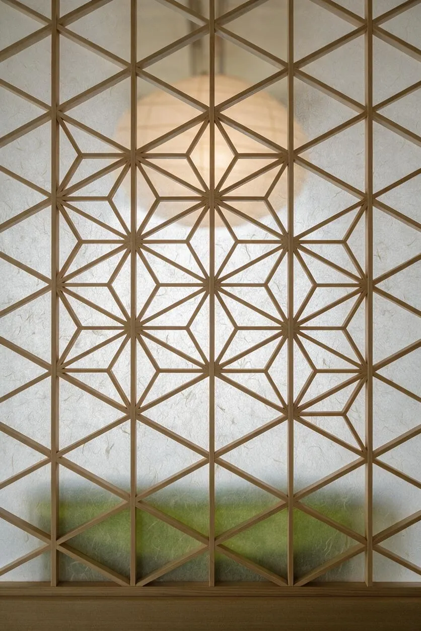 A Japanese Zen-style interior detail featuring a geometric wooden lattice screen over translucent rice paper. The pattern forms repeating diamond and star shapes, with soft light glowing behind the panel and a muted green band visible near the bottom. A wooden ledge runs along the base.