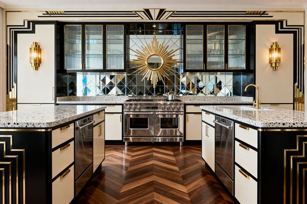 A glamorous Art Deco kitchen with two parallel islands in black and cream, each topped with speckled terrazzo and fitted with brass pulls. Centered on a stainless range, the back wall features diamond-pattern mirrored tiles and a gold sunburst, fluted-glass upper cabinets, tiered brass sconces, and bold stepped black-and-gold trim, all grounded by a warm walnut chevron wood floor in a perfectly symmetrical layout.