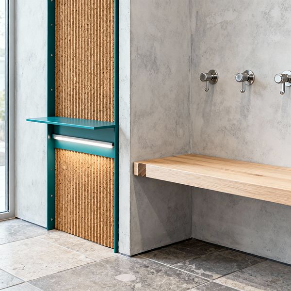 A modern mudroom features a floating light-oak bench fixed to pale gray concrete-render walls. Three brushed stainless steel hooks are mounted in a row above the bench. To the left, a vertical niche pairs a ribbed cork panel with teal powder-coated metal framing, a slim shelf, and an integrated LED strip. The floor is laid with large-format stone tiles in mixed gray and taupe tones, with a tall window at the edge bringing in daylight.