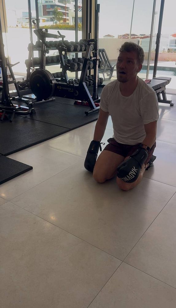 A man is kneeling on the gym floor, wearing boxing gloves and looking focused.